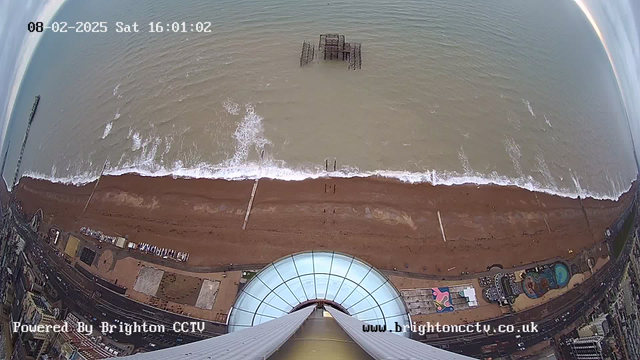 A view from above looking down at a beach and the sea. The sandy beach stretches across the bottom of the image, with a faint line of white waves lapping at the shore. In the center, there is a dilapidated pier partially submerged in the water. The seaside promenade is visible on the left, displaying a mix of buildings and structures. The sky is cloudy, and the time is displayed in the corner as 16:01 on February 8, 2025.