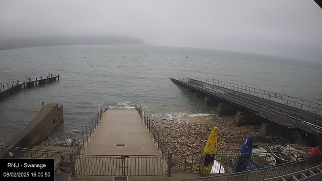 A view of a cloudy shoreline with a calm sea and mist in the distance. In the foreground, there is a set of concrete steps leading down to the water, enclosed by metal railings. To the right, there are several boats parked, including a yellow kayak. There are two piers extending into the water on the left, one with several posts and the other longer and flat. Rocks are visible along the water's edge, and the scene is under an overcast sky.