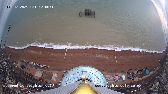 A high-angle view of a beach with a sandy shoreline. In the foreground, a circular glass structure appears to be part of a tall tower, possibly an observation point. In the background, the sea stretches out with gentle waves lapping against the shore. A weathered pier juts out into the water, and there are various structures, including a prominent amusement area with colorful designs, along the beach. The sky is bright, suggesting daytime with scattered clouds.