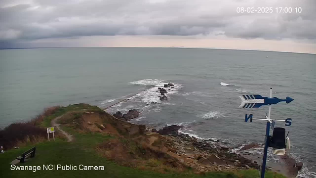 A coastal scene showing a rocky shoreline with waves crashing against the rocks. The sky is overcast with gray clouds, and the ocean appears calm, reflecting shades of blue and green. In the foreground, there is a grassy area leading to the edge of the cliff, with a wooden bench visible on the left side. A weather vane with directional indicators stands prominently on the right. There is also a sign partially visible near the path on the left. The date and time are displayed in the upper right corner.