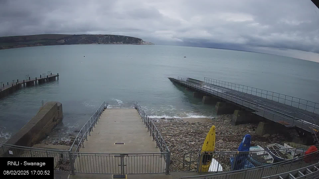 A view of a seaside area on a cloudy day. In the foreground, there is a concrete ramp leading down to the water, bordered by metal railings. On the right, two wooden piers extend into the calm sea; one is straight and the other slopes down. In the background, the faint outline of a chalk cliff or coastline is visible, shrouded by clouds. To the right of the ramp, there are several colorful kayaks (yellow, blue, and red) stored near a rocky shoreline. The water appears smooth, with a few small waves along the edge.