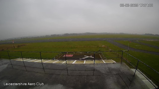 A cloudy sky is visible above a green field at an airfield. In the foreground, there is a railing overlooking a helicopter landing pad marked with a large white cross on the ground. The landing pad is surrounded by a fenced area, and further in the background, there is a runway with a visible marker at the edge. The landscape is flat with low hills in the distance. The scene is somewhat grey and overcast, indicating a dull day.