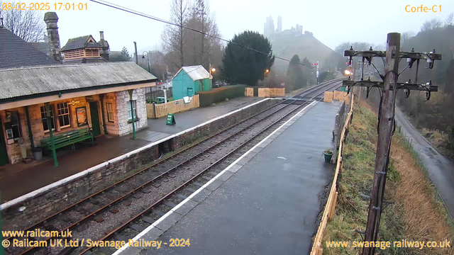 A partially foggy scene at a railway station. On the left, there is a stone building with a roof, featuring windows and a green bench outside. A green shed is visible behind the building. The railway tracks run horizontally across the image, with a neatly paved platform next to them. In the background, a hill rises, topped with remnants of a castle. A wooden fence and pathway are seen on the right side of the image. The ground is wet, suggesting recent rain, and the sky is overcast.