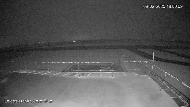 A dimly lit outdoor scene viewed from a webcam at the Leicestershire Aero Club. The image shows a flat surface, likely an airstrip, with faint outlines of markings on it. In the foreground, a railing runs along the edge of the platform, and beyond it, the landscape gradually fades into darkness. The time and date at the top right indicate it is 6:00 PM on February 8, 2025. There are no visible aircraft or people in the scene.