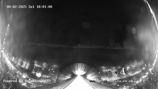 A black and white webcam image showing a coastal city at night. The foreground features a wide viewpoint of a public area with a promenade, where numerous lights and structures are visible. There are faint outlines of buildings and possibly rides or attractions, suggesting it is a lively area. The sky is dark, and the time stamp at the top reads "08-02-2025 Sat 18:01:00." Water can be seen in the background, reflecting some light. There are also some luminous dots in the air, possibly raindrops on the lens.