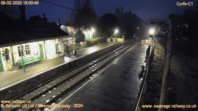 A dimly lit train station platform at dusk. A stone building with green trim is on the left, featuring windows and a bench. The ground is wet, reflecting light from nearby lamps. In the background, there are trees and a faint outline of a hill with structures. Train tracks run along the platform, leading into the distance. A small sign is visible on the platform, directing towards the exit. The scene is tranquil, with a few scattered lights illuminating the area.