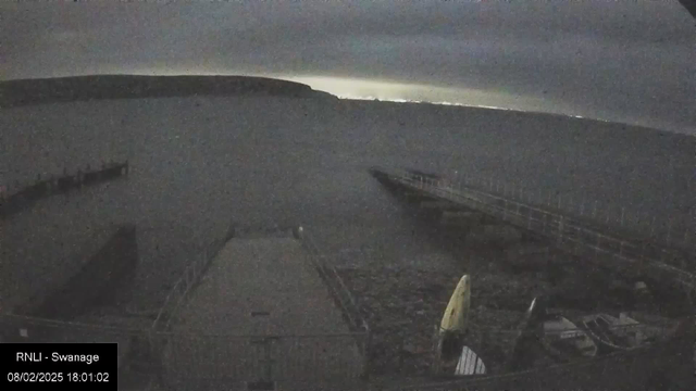 A dark waterfront scene at dusk, showing a wooden pier extending into the water on the right side. The water appears still and is reflecting some light, possibly from the horizon where a faint glow is visible in the distance. A few objects, including a small yellow sail and a white item, are partially visible on the left side of the image near the water's edge. The sky is overcast, creating a moody, subdued atmosphere.