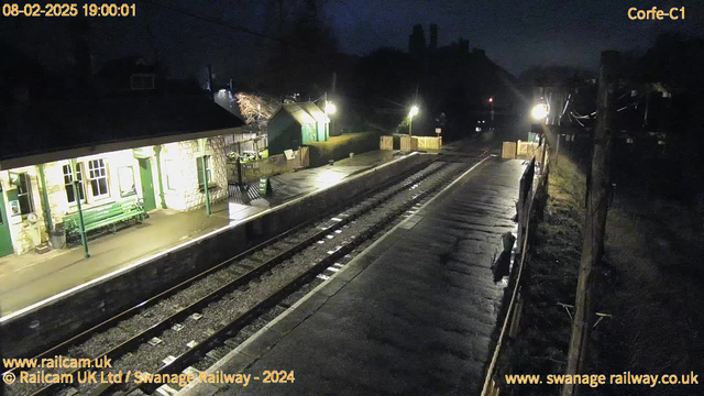 A dark, nighttime scene at a train station. The foreground features a platform with tracks running parallel, wet from the rain. To the left, a stone building with green trim houses benches and a sign. A few lights illuminate the area, casting reflections on the wet surface. Behind the building, a small green shed is visible. The background shows indistinct outlines of trees and hills. A wooden fence with a gate is in the distance, marking the edge of the station area.