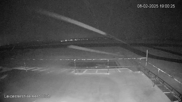 A dark, nighttime scene from a webcam at Leicestershire Aero Club. The foreground shows a flat surface with faint lines marking parking spaces, and a low railing runs along the edge. In the background, a dark sky is visible with hints of distant lights, possibly from an urban area. The overall ambiance is dim, with limited visibility of details. The timestamp in the corner indicates the date and time: 08-02-2025 at 19:00:25.