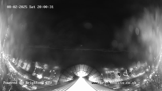 A black and white image from a webcam showing a cityscape at night. The image is dark with some bright spots representing city lights scattered across the lower half. The upper portion features a cloudy sky, and in the center, there is a bright area suggesting a light source, likely a building or structure. The date and time are displayed in the upper left corner. The scene has a sense of depth with structures extending toward the horizon.