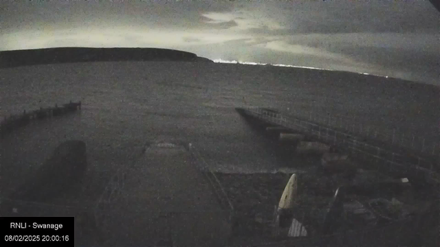 A dimly lit view of a body of water under a cloudy sky, with a distant shoreline visible on the left. In the foreground, a wooden pier extends from the bottom right into the water. Various objects, possibly boats or equipment, are scattered near the pier. The overall atmosphere is serene and somewhat mysterious with low visibility due to the twilight conditions.