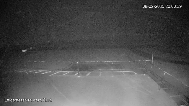 A dark image depicting a scene from a webcam at Leicestershire Aero Club. The view shows a dimly lit area with a low fence and several lines of what appears to be parking spaces, possibly for aircraft. The date and time displayed in the corner indicate it is 8th February 2025, at 20:00:39. The atmosphere feels quiet and still, with no visible activity.