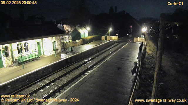 A nighttime view of a railway station with dim lighting. Two platforms are visible, one with a stone building that has a green roof and large windows. A green bench is located on the platform near the building. The opposing platform has a small wooden shed, and a white sign partially obscured that appears to say "WAY OUT." The area around the tracks is wet, reflecting light from the nearby lamps. In the background, there are silhouettes of trees and structures against the dark sky.