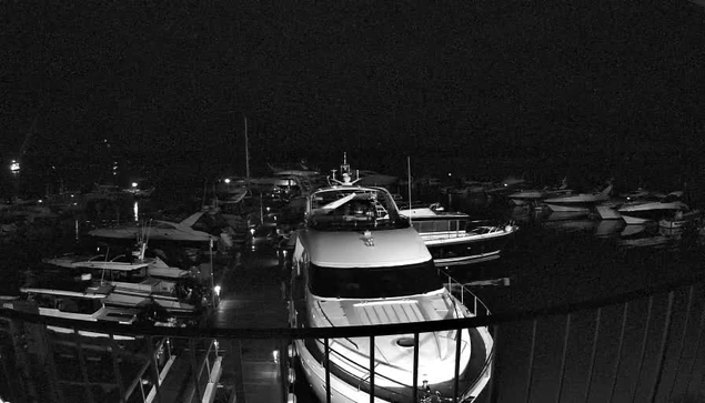 A dimly lit marina scene at night, featuring numerous boats docked in the water. In the foreground, a large white yacht is prominent, while smaller boats are scattered around. The atmosphere is calm, with faint reflections of lights from the boats shimmering on the water's surface. The area is mostly dark with some illuminated spots, contributing to a peaceful nighttime ambiance.