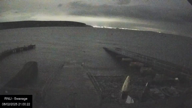 A dark scene featuring water and a distant shoreline under a cloudy sky. A wooden pier extends into the water from the foreground, partially illuminated by faint lights in the distance. The overall mood is tranquil with muted colors, suggesting dusk or nighttime conditions.