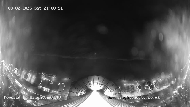 A dark webcam image showing a city scene at night, with a large illuminated area in the center suggesting a building or structure. Surrounding the bright area are blurred outlines of streets and possibly vehicles. Reflections and light spots appear, indicating moisture on the lens. The date and time display is visible in the top left corner, while a logo is present in the bottom left. The overall scene is mostly in shades of black and gray.