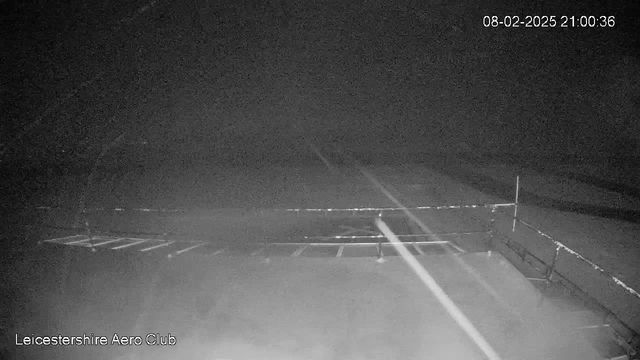 A dark and foggy scene is visible, with faint outlines of a runway and a barrier at the Leicestershire Aero Club. There are indistinct markings on the ground that suggest parking spaces. The image appears to be taken at night, with very low visibility. A timestamp in the top right corner indicates the date and time as February 8, 2025, at 21:00:36.