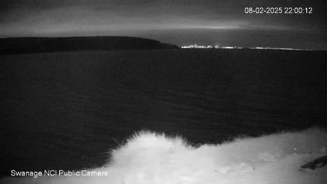 A dark, nighttime scene of a coastline. The water is visible, reflecting dim light, with gentle waves lapping at the shore. In the distance, faint lights from a town or city can be seen along the horizon. The sky is overcast, adding to the overall dark ambiance of the image. The date and time are displayed in the upper right corner.