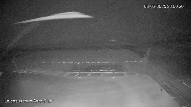 Image of a dark, nighttime scene captured from a webcam at Leicestershire Aero Club. The view includes a flat, open area with faint lines that may represent a runway or taxiway surface. There are faint lights in the distance, possibly indicating parking spaces. The date and time in the corner indicate February 8, 2025, at 22:00:20. The overall image lacks color and detail, contributing to the nighttime ambiance.