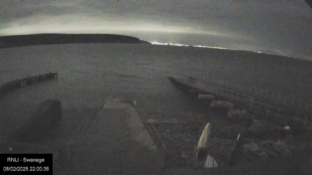 A dark shoreline scene with a body of water extending into the distance. In the foreground, a partially visible dock with wooden beams and a small white boat is seen resting on the shore. The sky is overcast, with faint hints of light on the horizon suggesting distant lights or reflections. The landscape gradually rises into a dark silhouette of land on the left side of the image. The overall mood is calm and quiet, with subdued colors and minimal visibility.