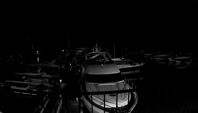 A nighttime view of a marina with several boats docked. In the foreground, a large silver boat is prominent, with its shape and features slightly illuminated. Surrounding it are various other boats, mostly dark, blending into the night. A railing is visible in the foreground, creating a sense of depth. The water reflects faint lights from the marina, hinting at the presence of additional boats in the background. Overall, the scene is predominantly dark, with limited visibility.