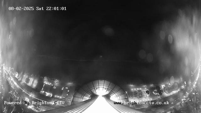 A black-and-white webcam image taken at night showing a view from a high point. The bottom of the image features a wide structure partially illuminated, with a downward slope leading toward dark areas on either side. Below, city lights are faintly visible, with a curved shape suggesting the presence of buildings. The date and time in the upper left corner indicate it is February 8, 2025, at 10:01 PM. The bottom includes text indicating the image is powered by Brighton CCTV. The overall atmosphere is moody, with a sense of height and distance.