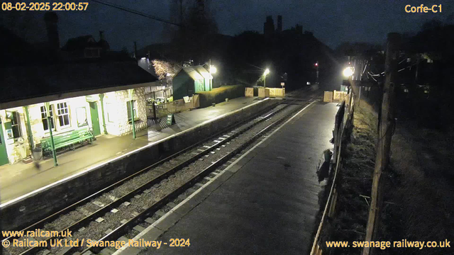 A dimly lit railway station scene at night, featuring a platform with a stone building on the left and wooden fences on both sides. There are two green benches in front of the building, which has illuminated windows. The ground is wet, reflecting light from nearby lamps. In the distance, train tracks run parallel to the platform, leading out of the frame. Various items, including a sign indicating "WAY OUT," are scattered around the area.