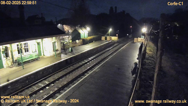 A nighttime view of a railway station platform. The platform is empty and wet, reflecting light. On the left, there is a stone building with green trim and a green bench outside. There are minimal lights illuminating the area, one near the platform and another closer to the building. In the background, there are trees and a slight incline leading to a structure or hill. A small sign stands on the pavement reading "WAY OUT." The image captures a serene, quiet atmosphere typical of a late hour at a train station.