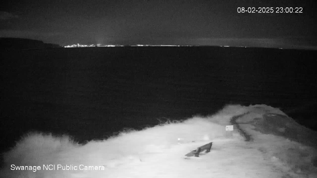 A nighttime view of the ocean with dark water reflecting a range of lights from a distant shore. In the foreground, there are gentle waves crashing against a rocky seaside. The coastline is silhouetted against the dark sky, and a small wooden structure is partially visible near the water. The date and time are displayed in the corner.