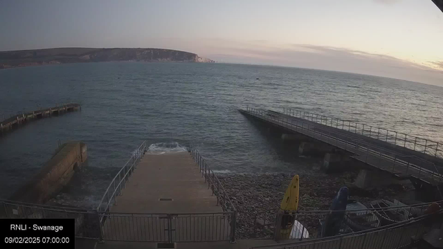 A coastal scene showing the sea at dawn. In the foreground, there is a wooden walkway leading down to a concrete ramp that extends into the water. To the left, a stone pier juts out into the sea. Multiple colors of kayaks are visible on the shore. The water is calm with gentle waves, and the sky is light blue with soft clouds, indicating early morning light. In the background, a hilly coastline can be seen.