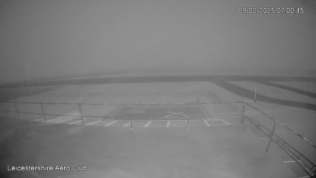 A grayscale image of a foggy scene at an airstrip. The foreground shows a flat area with a grid pattern marked on the ground, possibly indicating parking spaces. There is a low fence along the edge of the area, and in the background, the horizon is almost indistinguishable due to the fog. The time and date are displayed in the corner, indicating the image was taken early in the morning.