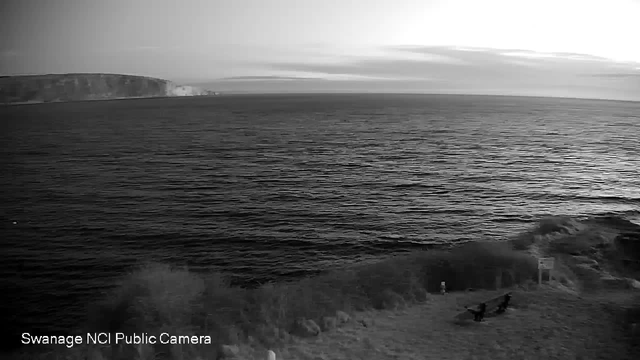 A black and white image of a coastline at dusk. The foreground shows a grassy area with silhouettes of two people sitting on benches, facing the water. The middle ground features calm ocean waters reflecting the light, with gentle waves. In the background, a cliff rises sharply from the sea under a cloudy sky, with dark shadows creating a sense of depth. The overall mood is tranquil and serene.