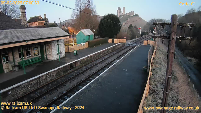 The image shows a train station setting in the early morning. On the left, there is a stone-built station building with a sloped roof and green benches outside. Next to the station, there is a small green wooden shed. A narrow path runs along the platform next to two railway tracks, which lead off into the distance on the right. In the background, a hill features the ruins of a castle, and there are some trees scattered around the area. The scene is illuminated by soft morning light, creating a tranquil atmosphere.