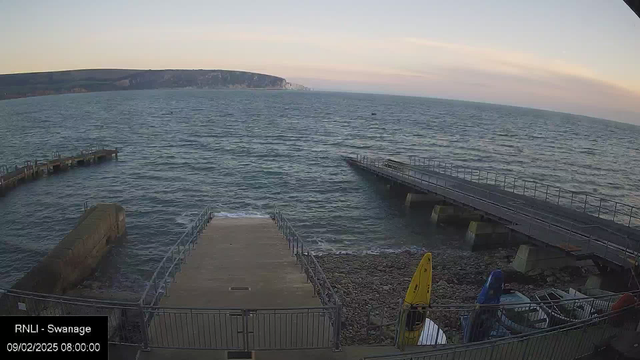 A view of a calm sea with gentle waves, under a clear sky with soft colors of dawn. In the foreground, there is a wooden jetty extending into the water, accompanied by a rocky shoreline on the left. To the right, a second jetty juts out, and several colorful kayaks are visible on the rocky ground. The scene is quiet and serene, capturing the beauty of a coastal landscape.