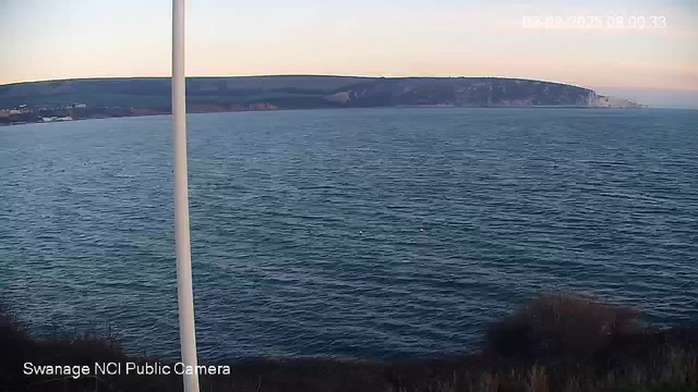 The image shows a view of the ocean from a high vantage point. The surface of the water is slightly rippled, reflecting a subtle blend of blue and green hues. In the background, there are rocky cliffs and green hills. A white pole stands on the left side of the image. The sky above is light with a hint of pink, indicating early morning or sunset. Near the water, a few buoys can be seen floating.