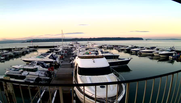 A marina at sunset featuring numerous boats docked in calm water. In the foreground, a large white boat with a cabin sits moored. Surrounding it are smaller vessels of various sizes and colors, all reflecting in the water. The sky is painted in soft hues of orange and pink, creating a serene atmosphere. In the background, a distant shoreline is visible with a gentle slope and trees silhouetted against the sky.