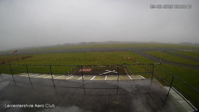 A view from a webcam positioned on a building overlooking a large grassy area. The scene is foggy and overcast, with low visibility. In the foreground, there is a railing, and a visible paved area marked with an "X". In the background, there are wide, open fields and a runway that stretches off into the distance. A windsock is visible on the left side, indicating light wind conditions. The time and date in the corner shows it is early morning on February 9, 2025.