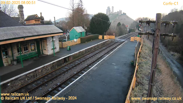 A railway station scene is depicted, featuring a stone building with a sloped roof, a green bench in front, and a wooden fence to the right. Railroad tracks stretch along the foreground, leading to the background where a hill topped with ruins is visible. To the left, a green shed stands and there are trees around the area. The sky is light, indicating morning, with utility poles lining the tracks.