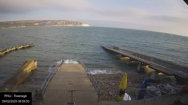 A view of the seaside with a concrete ramp leading down to the water. On the left side, a wooden pier extends out into the sea, while more wooden decking can be seen farther along the shore. Kayaks in yellow and blue are stored nearby on the rocky beach, and gentle waves can be seen lapping at the shore. The background features green hills and cliffs under a partly cloudy sky. The scene is tranquil and suggests early morning light.