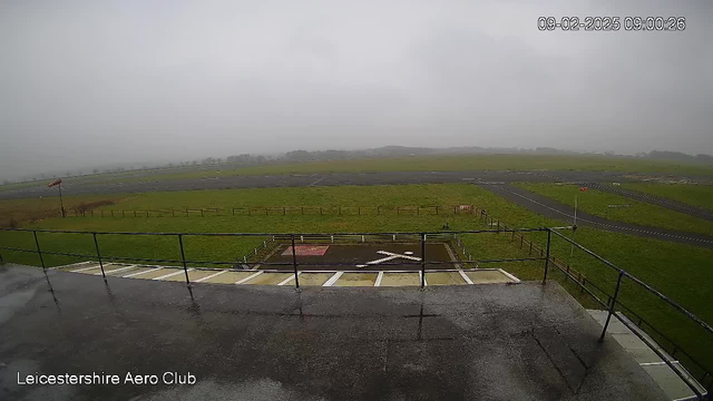 A view from a balcony overlooking a large open field and runway, with gray, cloudy skies and low visibility. The ground appears wet, possibly from rain. A red wind sock is visible to the left, indicating wind direction. In the foreground, there's a marked area with a white 'X'. The surrounding area is grassy, with a wooden fence bordering the field and a road running alongside the runway. The image timestamp reads 09:00:26 on February 8, 2025.