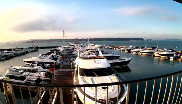 A marina scene with several boats docked in the water. In the foreground, a large white yacht is prominently positioned, surrounded by smaller boats of various sizes and designs. The water is calm, reflecting the sky, which is partly cloudy with a hint of sunlight. In the background, hills are visible along the shoreline. The image conveys a serene setting with a focus on leisure and boating activities.