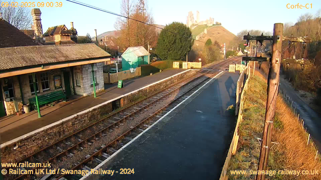 A sunny day at a train station with a stone building on the left featuring a sloped roof and a covered area with benches. In the foreground, there's a platform with a green sign indicating “OUT.” The railway tracks are visible extending into the distance. To the right, there’s a wooden utility pole with power lines. In the background, a hill rises with a castle-like structure on top, surrounded by trees. The scene conveys a peaceful, rural atmosphere.