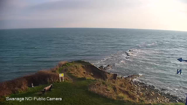 A scenic view of the sea from a cliff edge, with gentle waves lapping at the rocky shoreline. In the foreground, there is a grassy area featuring a bench and a warning sign. The background shows the expansive ocean under a slightly cloudy sky with the sun shining, casting a soft light over the water.