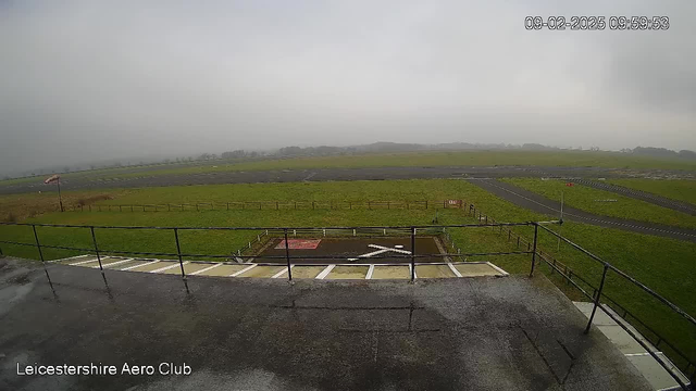 A cloudy, overcast sky hovers over an open field that connects to an airstrip. In the foreground, there is a railing with a wet surface, suggesting recent rain. To the left, a helipad marked with a white "X" is visible, and further back, a grassy area leads to a paved runway. The scene is quiet and desolate, with no visible aircraft or people. A flagpole can be seen with a flag gently flapping in the wind. The timestamp in the corner reads 09:59:53 on February 9, 2025.