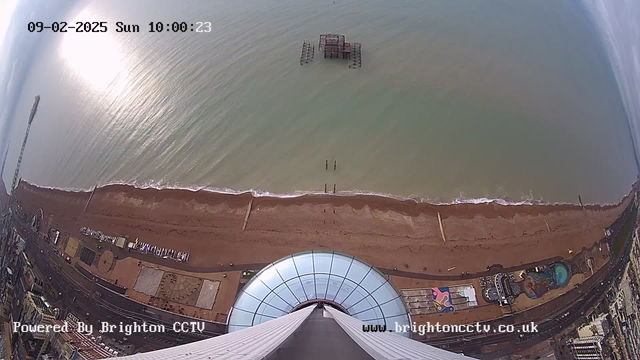 Aerial view of a beach with sand and intermittent waves. The shoreline is lined with wooden posts and a derelict structure is visible in the water. On land, there are amusement rides and various attractions. The top of a circular structure is seen in the foreground, and the image has a timestamp showing the date and time.