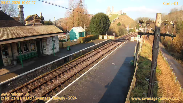 A railway station scene is depicted, featuring a platform on the left with a stone building that has a sloped roof, a green bench, and several trees in the background. The railway tracks run parallel to the platform, leading into the distance. In the background, a castle can be seen atop a hill, surrounded by trees and a blue sky. A utility pole with wires is situated on the right side of the image, and a sign labeled "WAY OUT" is visible near the platform edge. The scene is well-lit, indicating a clear day.