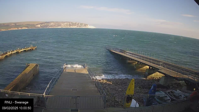 A view of a coastline with a gentle sea and clear skies. In the foreground, there are two piers extending over the water, with one pier leading directly from a concrete ramp. The ramp has a railing along its side, and waves are gently lapping against the concrete. To the right, there are several colorful kayaks stored on a rocky beach area, with a few boats partially visible behind them. In the distance, grassy hills rise up, leading to white chalk cliffs under the blue sky.