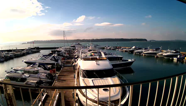 A busy marina scene shows numerous boats lined up at dock. In the foreground, several yachts are moored, with some featuring canopies and bright colors. The water is calm and reflects the sky, which is partially cloudy with patches of blue. In the background, additional boats are anchored further out, and green hills are visible along the horizon. A pier extends into the water, adding depth to the scene.