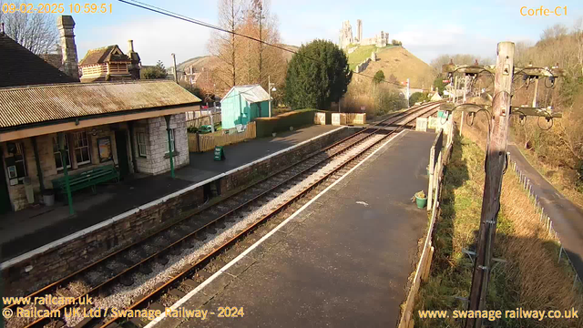 A railway station platform is shown, with a building on the left featuring a roof and stone walls. To the right, there are railway tracks leading into the distance. A green wooden bench is visible on the platform, along with a blue shed and a few trees. In the background, a hill rises with ruins or a castle on top, under a clear blue sky.