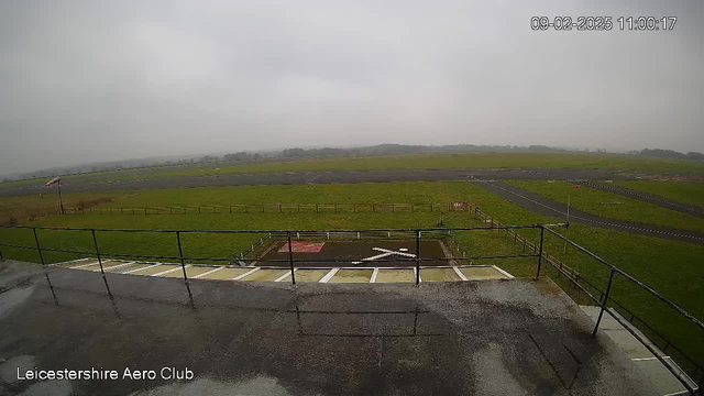 A cloudy sky looms over a rural airfield. In the foreground, a balcony railing frames the view. Below, the airfield features a grassy area bordered by a wooden fence, with a runway made of asphalt visible in the background. Various markings on the ground indicate different areas, including a red square with a white cross. The scene appears damp, suggesting recent rain.